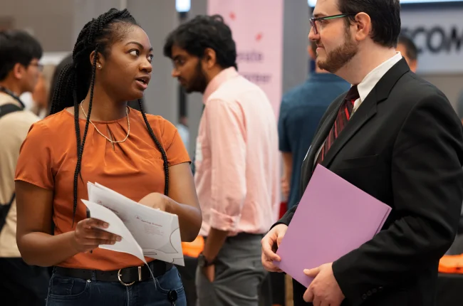 A student points at papers in her hand as she speaks with a man at a networking event.
