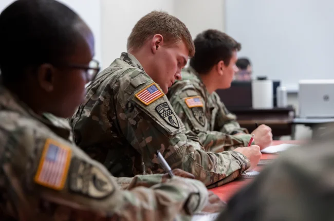 Three students in ROTC uniforms sit at tables taking notes during class.