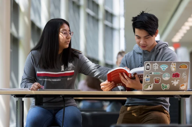 Two students sit at a table, one pointed at a book in the other’s hand.