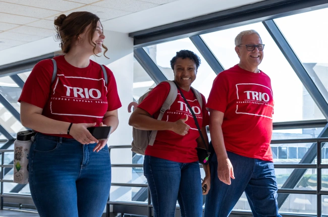 Three people wearing red TRIO Student Support Services shirts walk through a campus building lit by modern, angled windows.