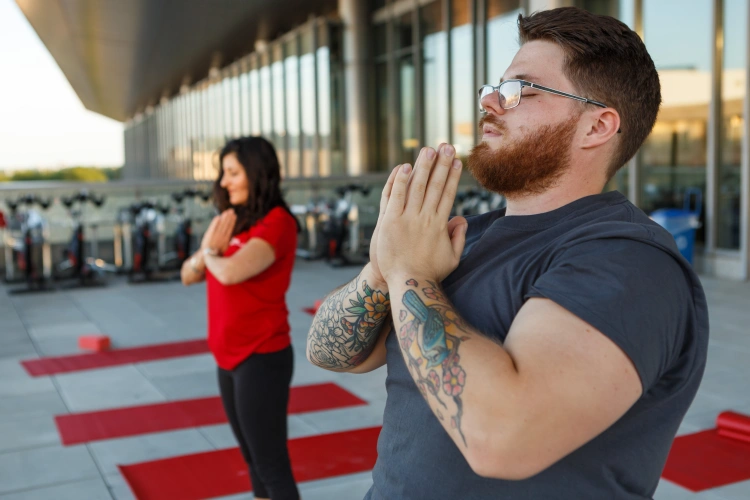 Two people stand on yoga mats laid out on the deck of the campus center, eyes closed and hands palm to palm in front of them.