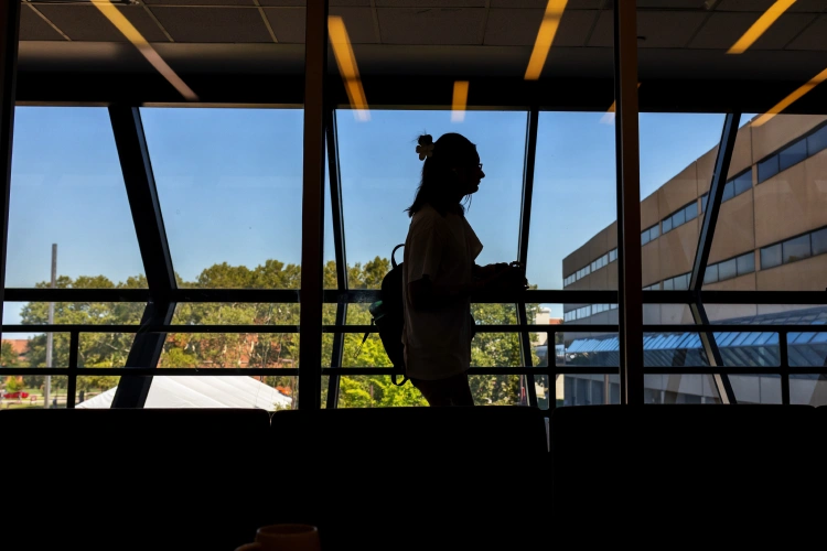 A student in silhouette walks past a bank of angled windows overlooking campus.