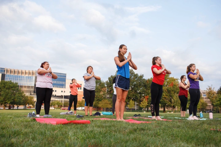 A group or about ten people stand on mats, hands pressed together in front of them, for an outdoor yoga class on a green lawn.